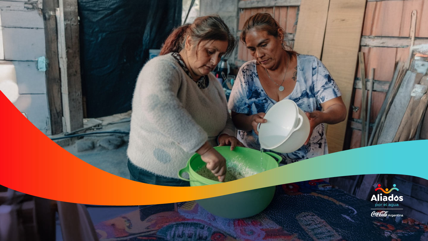 Dos mujeres de mediana edad, preparan alimentos en un entorno doméstico rústico. Una mezcla en un bowl verde mientras la otra vierte agua de un recipiente blanco. En la esquina inferior derecha, el logo 'Aliados por el agua' de Coca-Cola Argentina.