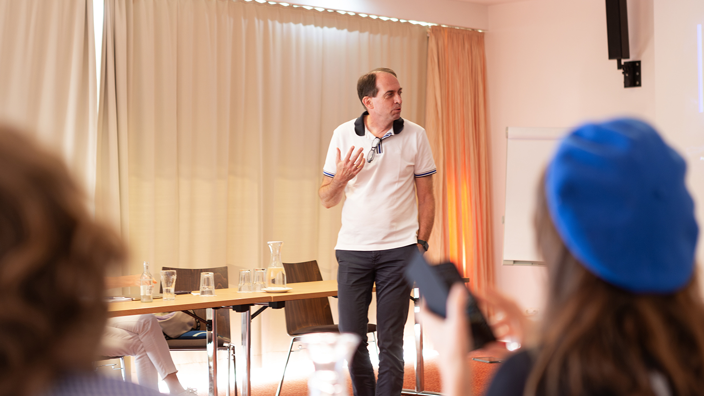 Ein Mann im weißen Polo-Shirt steht vor einer Gruppe und spricht. Im Vordergrund sind unscharf Teilnehmende zu sehen, auf dem Tisch davor stehen Gläser und Wasserflaschen.
