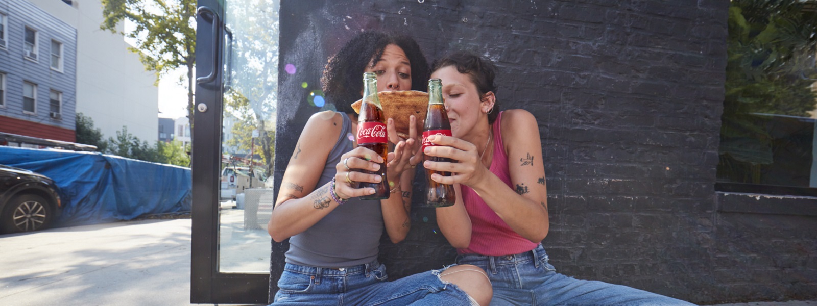 Dvije mlade nasmijane osobe, sjede na podu, jedu pizzu i piju Coca-Colu iz staklenih bočica