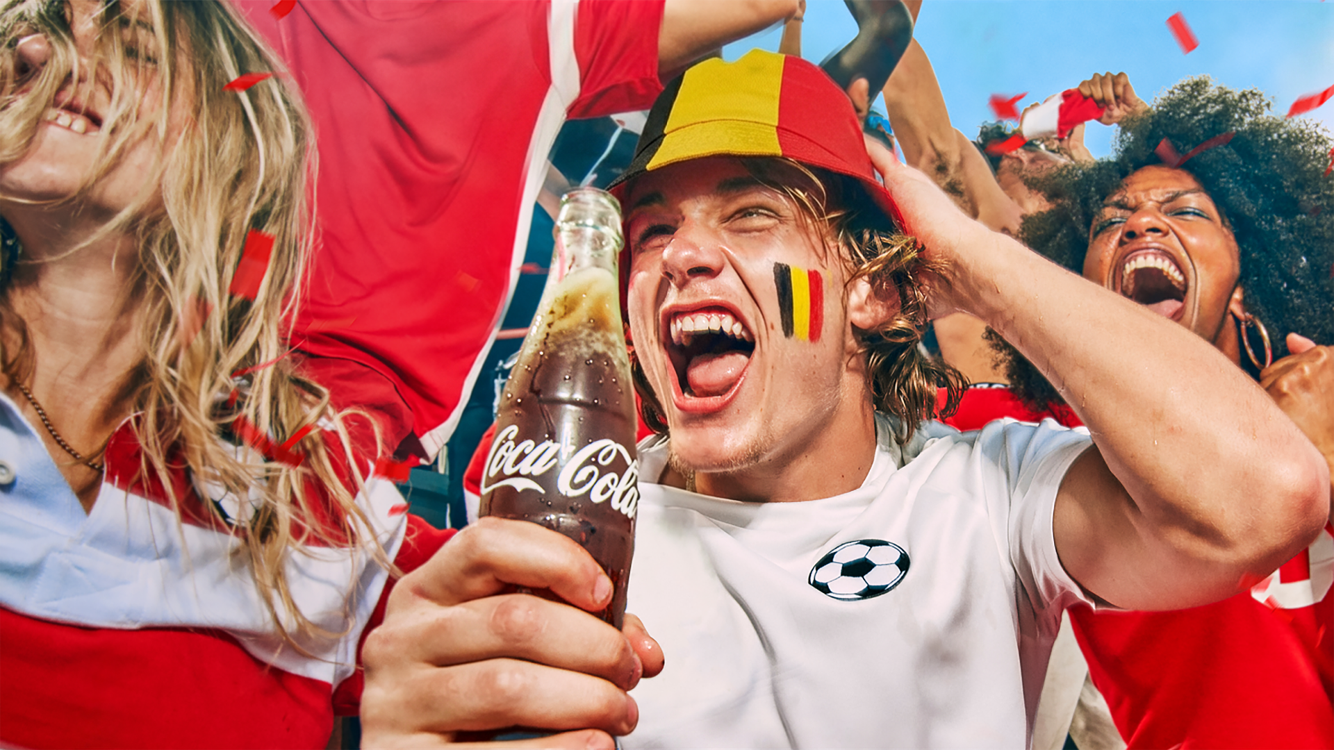 Groupe festif de supporters vêtus de rouge et blanc en train de célébrer ; au premier plan, une bouteille de Coca‑Cola bien fraîche est levée tandis que des confettis tombent, évoquant une ambiance estivale et footballistique.