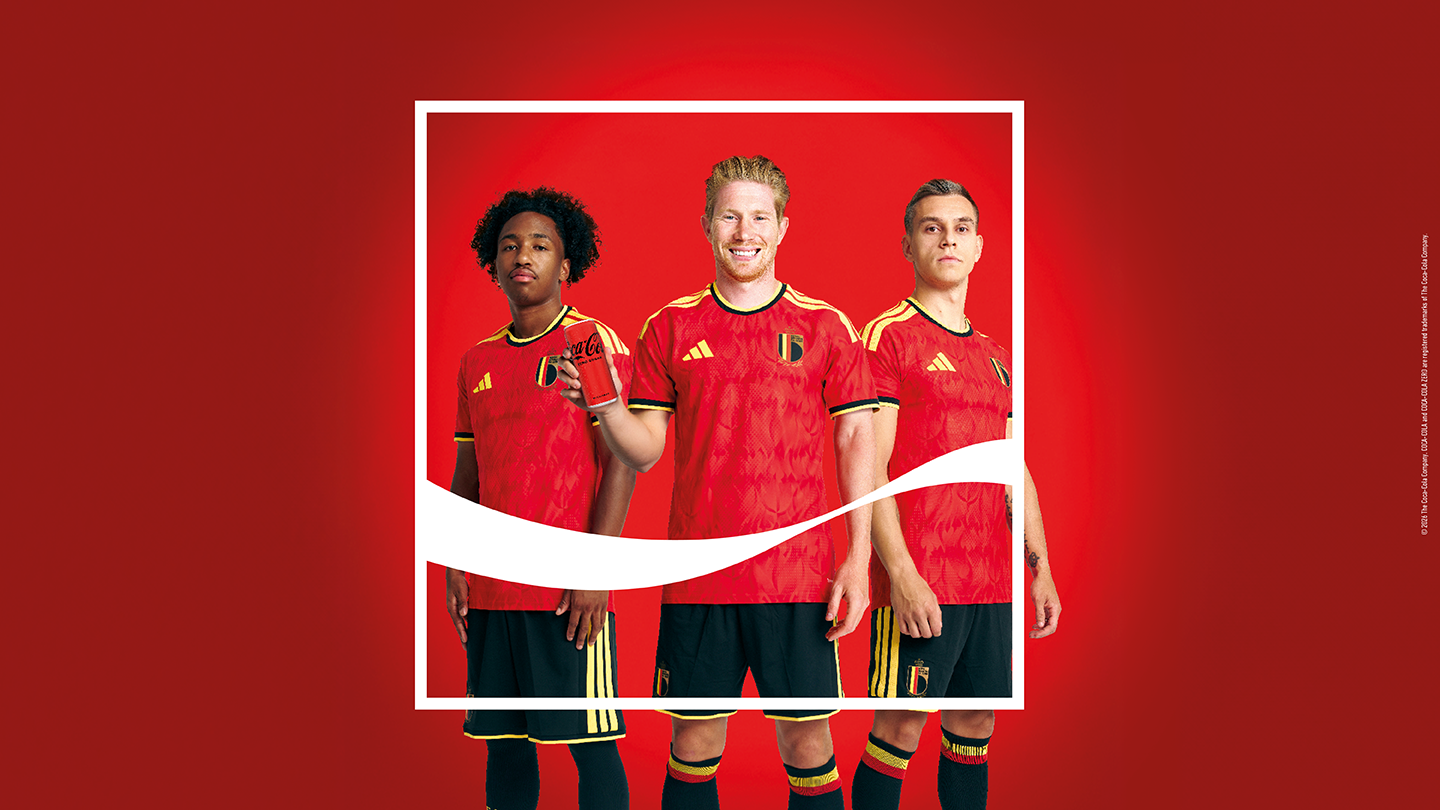 Drie voetballers in rode wedstrijdshirts met zwarte en gele accenten staan naast elkaar tegen een rode achtergrond, omkaderd door een wit kader met een golvende lijn; de middelste persoon houdt een rood Coca‑Cola‑blikje vast.