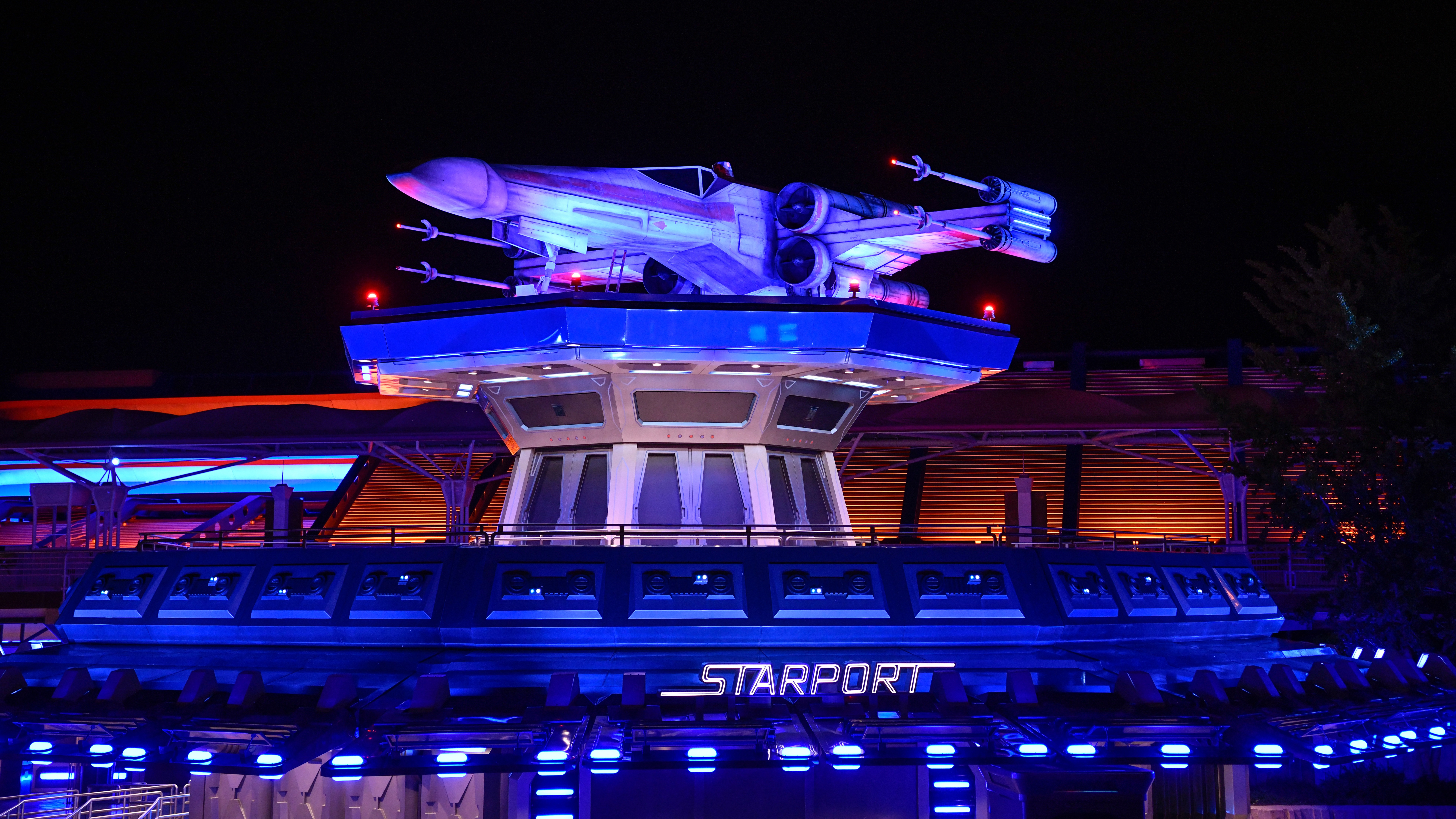 X‑wing ruimteschip tentoongesteld boven de ingang van de Starport‑attractie in Disneyland Paris, verlicht met blauwe en paarse lichten tegen een nachtelijke hemel.