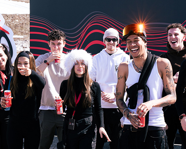 Um grupo de jovens sorridentes desfrutando de Coca-Cola em um ambiente de inverno, com roupas quentes e óculos de sol.