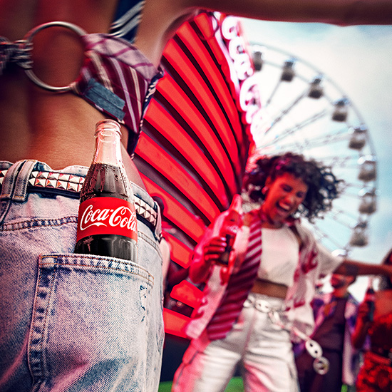 Uma garrafa de Coca-Cola no bolso de uma calça jeans com uma cena de festival alegre ao fundo.