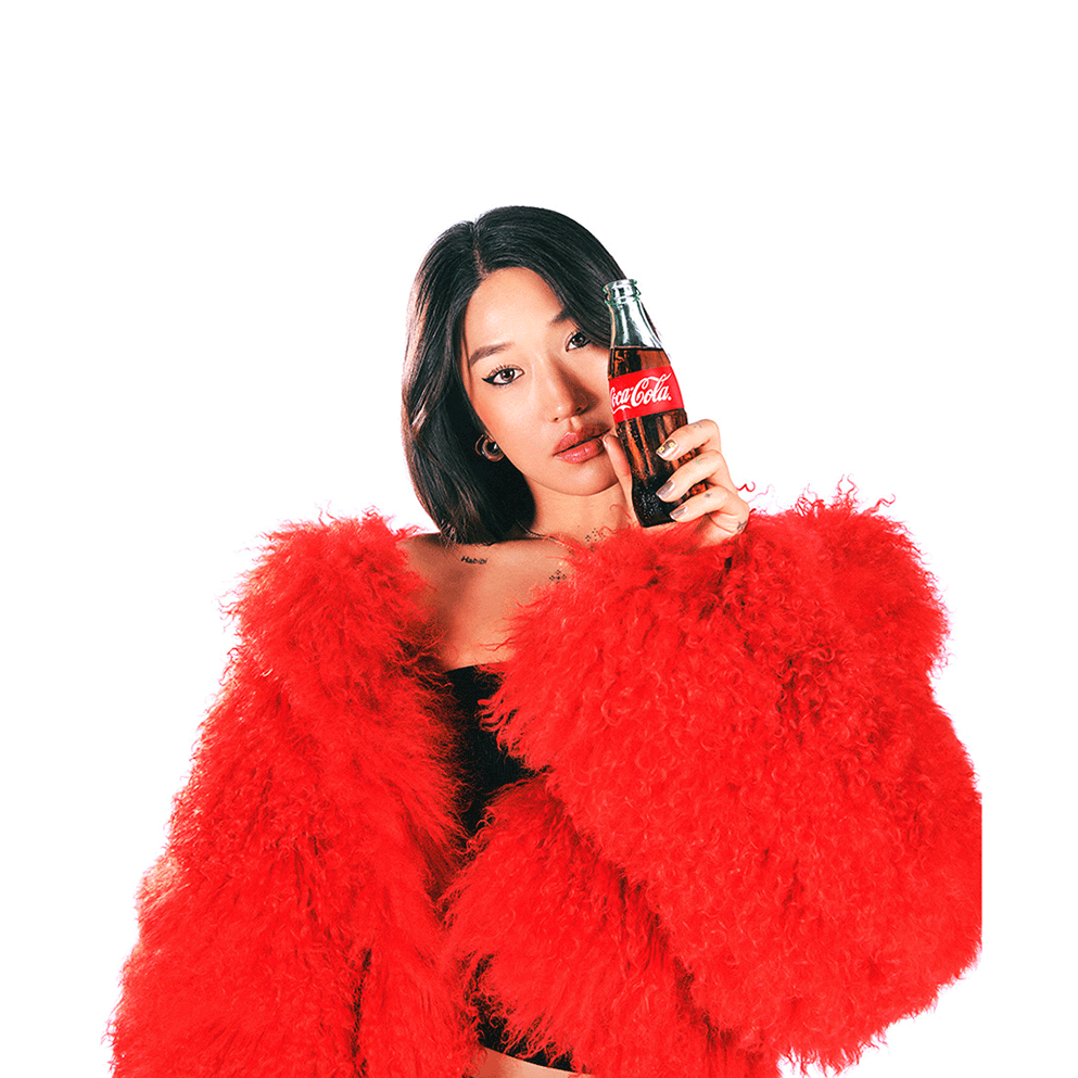 Peggy Gou, uma jovem de cabelo escuro e longo, veste um casaco de pelúcia vermelho brilhante e segura uma garrafa de vidro de Coca-Cola perto do rosto, olhando para a câmera com uma expressão tranquila.