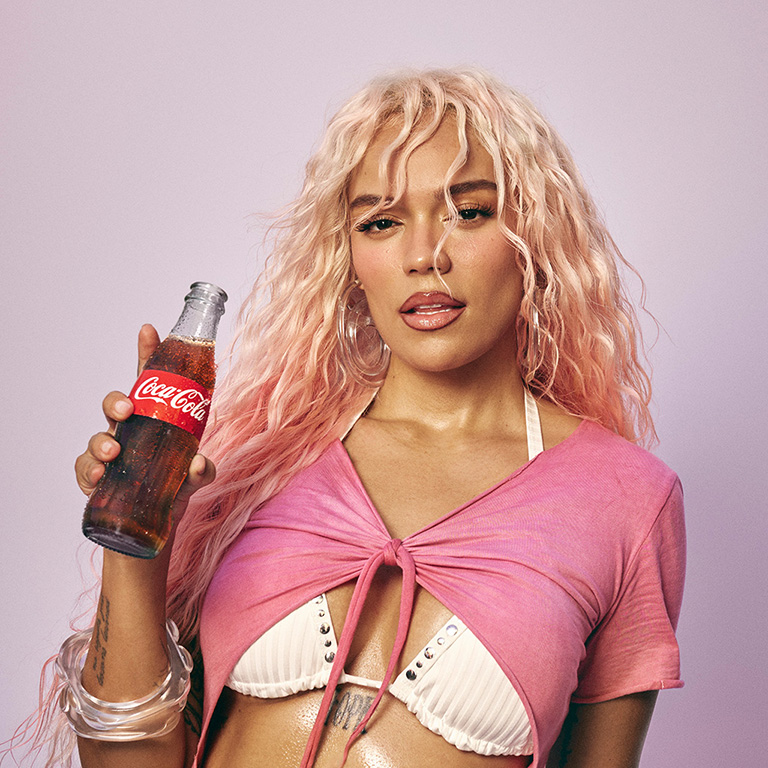 Karol G posando com uma garrafa de Coca-Cola em frente a um fundo neutro.