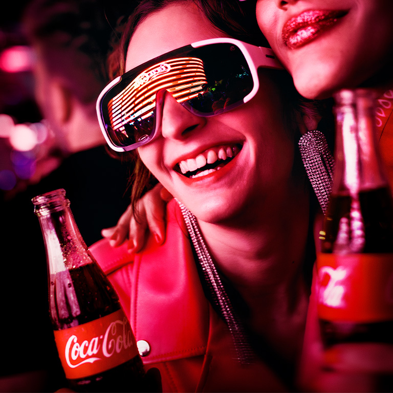 Pessoa com óculos futuristas e uma garrafa de Coca-Cola em um ambiente de festa.