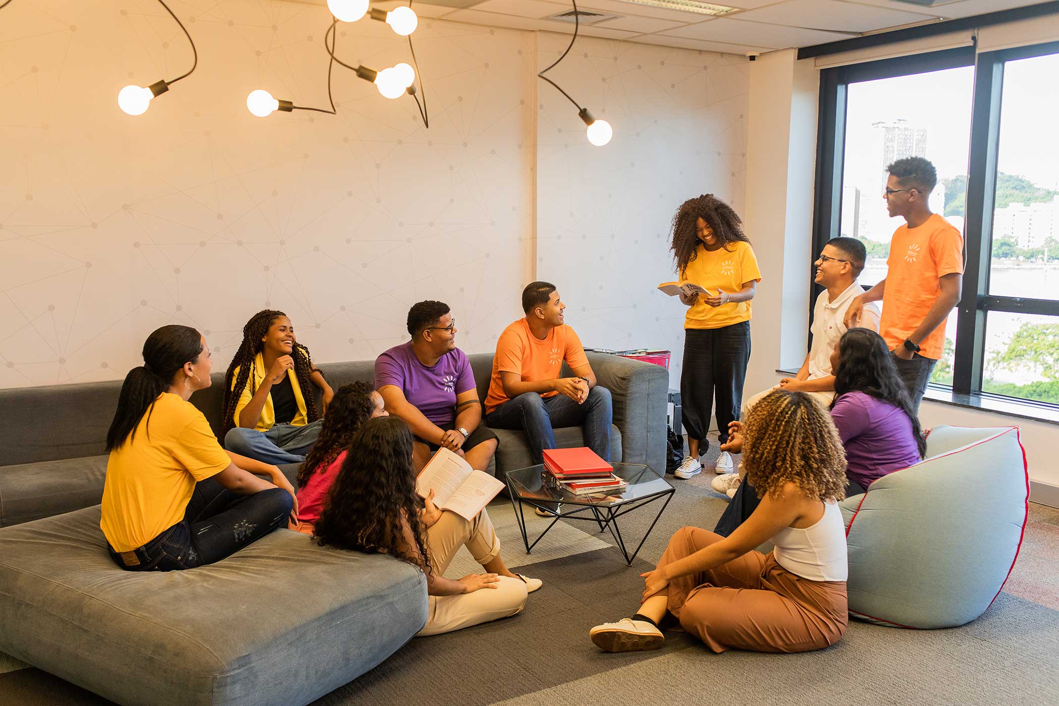 grupo de pessoas reunidas em uma sala, algumas sentadas em um sofá e outras em pé. Eles têm camisetas amarelas, roxas e brancas.