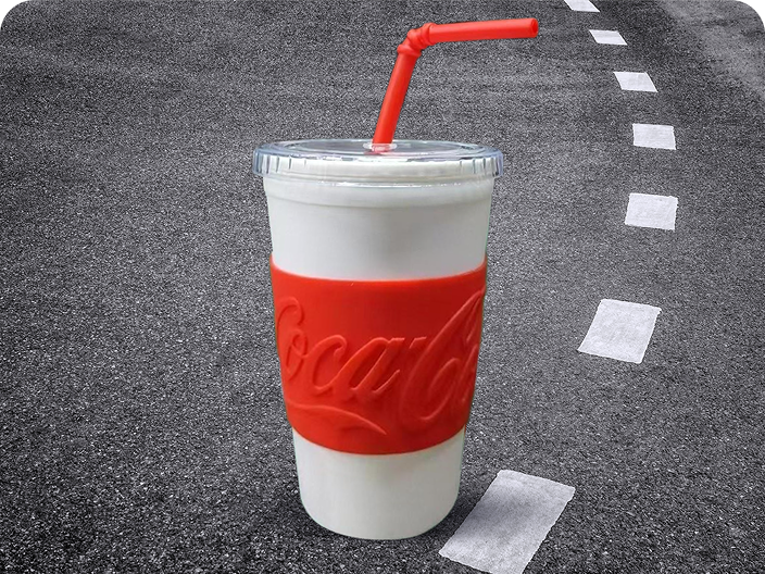 Un gobelet blanc avec un manchon et une paille rouges Coca-Cola, un prix du concours Voyage en voiture, sur un fond d'asphalte.