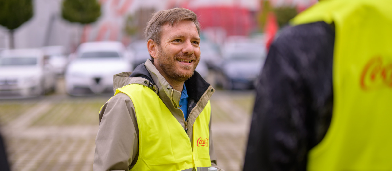 Ein Mann mit einer Coca-Cola Warnweste hält eine leere PET-Flasche in der Hand und unterhält sich mit einem anderen Mann.