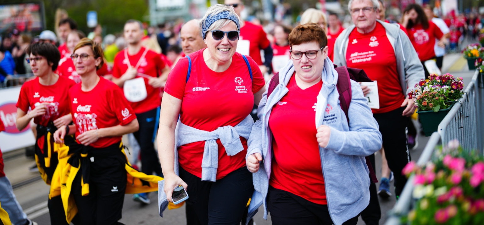 Zwei Läuferinnen am Coca-Cola Inclusion Run 2025 in Wien schauen in die Kamera, während hinter ihnen weitere Läuferinnen die Strecke bestreiten.