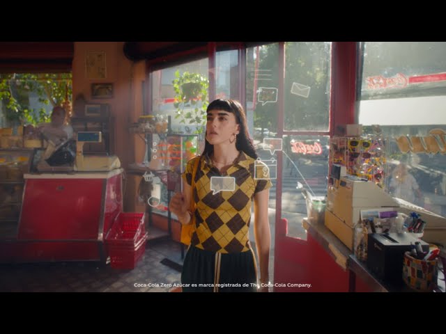 Joven de pie en el interior de una tienda, sosteniendo una botella de Coca‑Cola Zero Azúcar, con decoración y luz natural entrando por las ventanas.