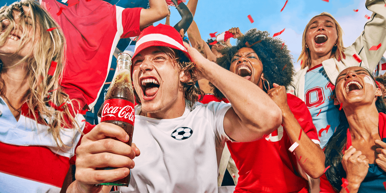 En gruppe jublende fans i rødt og hvidt fejrer sammen udendørs. I forgrunden holder en person en flaske Coca-Cola, mens konfetti flyver i luften og stemningen er festlig og energisk.