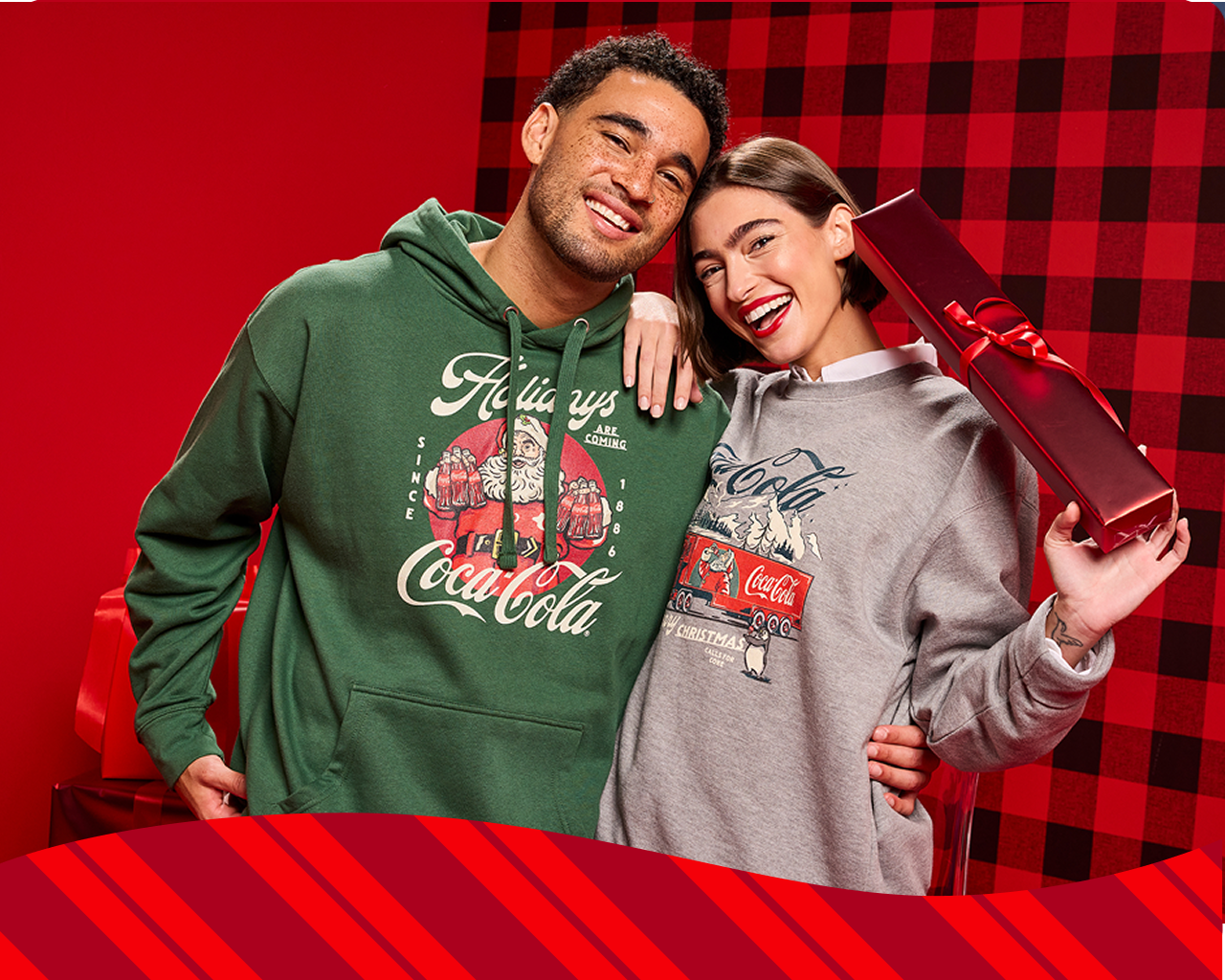 Paar hält ein verpacktes Geschenk in Coca‑Cola‑Weihnachts-Sweatshirts mit Santa‑Motiven.  Santa trinkt Coca‑Cola aus einer Glasflasche vor einem roten Punktmuster-Hintergrund.