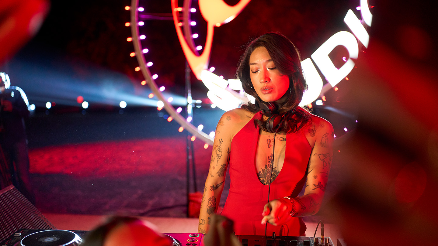 Peggy Gou en un evento de Coke Studio, tocando en un escenario con iluminación roja y el logo de fondo. Lleva un vestido rojo y audífonos colgando del cuello, en un entorno de fiesta.