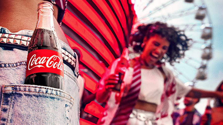 Una botella de Coca-Cola en el bolsillo de un pantalón de mezclilla, con una escena alegre de festival en el fondo.