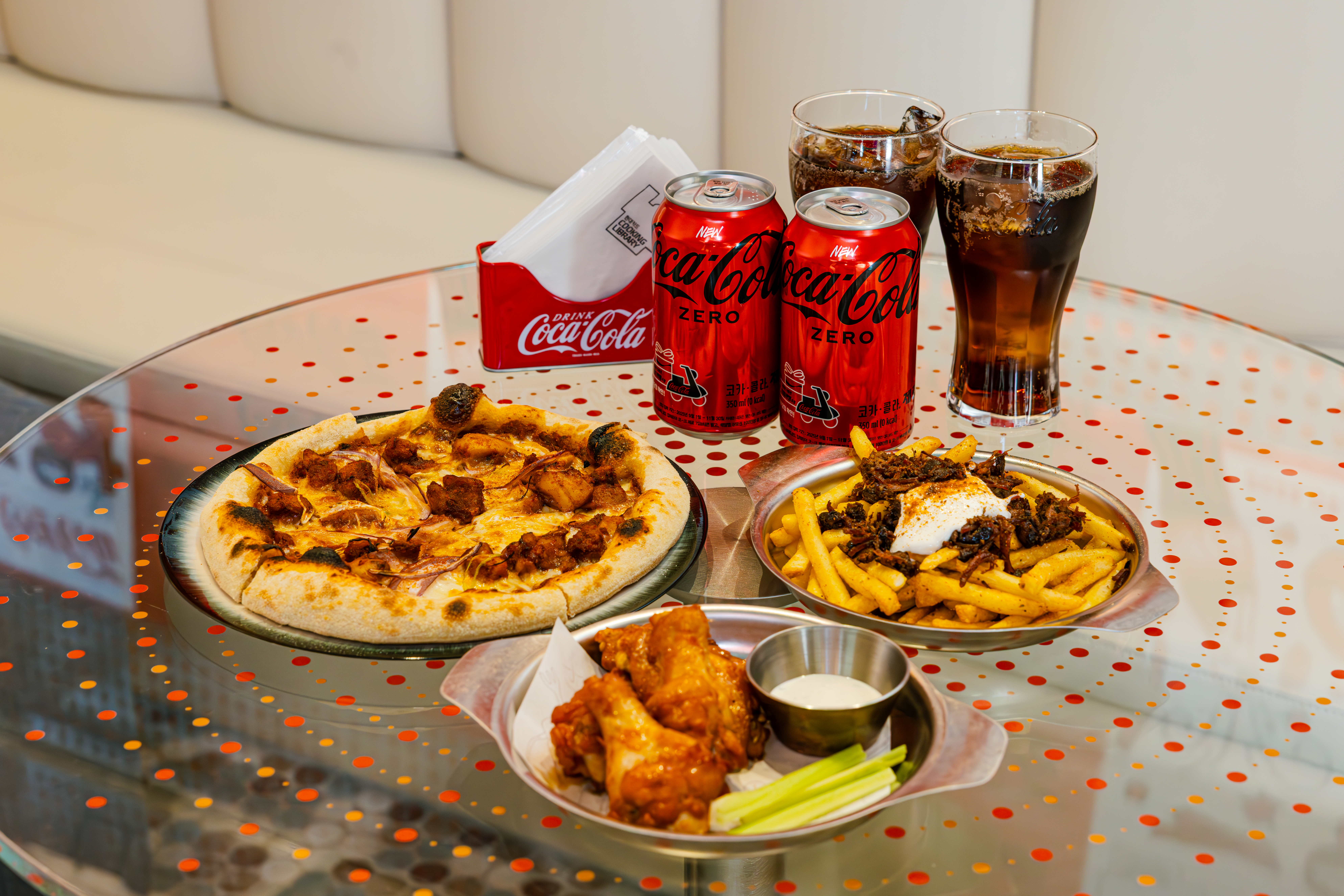 1층 델리에서 즐길 수 있는 특별한 코카-콜라 콤보메뉴 3종. 치폴레 치킨 피자와 비프 베이컨 감자튀김, 그리고 버팔로윙, 그리고 코카-콜라 제로가 먹음직스럽게 테이블 위에 올라와 있다.