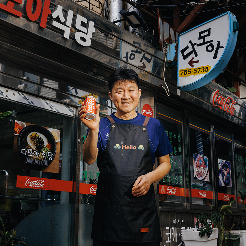 신흥시장 다모아식당 유재윤 사장이 식당 앞에서 코카-콜라 캔을 들고 환하게 웃음 짓고 있는 모습.