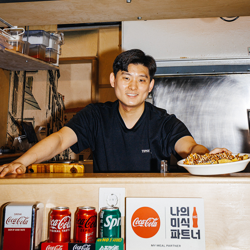 신흥시장 팁시타코 유영섭 사장이 주방 카운터 뒤에서 미소 지으며, 직접 만든 멕시칸 요리를 접시에 담아 내미는 모습.