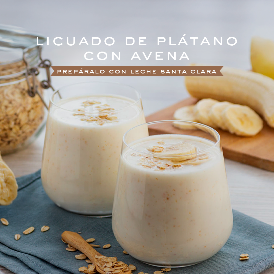 Dos vasos de licuado de plátano con avena, decorados con rodajas de plátano y avena espolvoreada, sobre una tela azul. Al fondo, se ve un frasco de avena, un plátano entero y rodajas de plátano en una tabla de madera. En la parte superior de la imagen, un texto dice "LICUADO DE PLÁTANO CON AVENA" y debajo "PREPÁRALO CON LECHE SANTA CLARA".