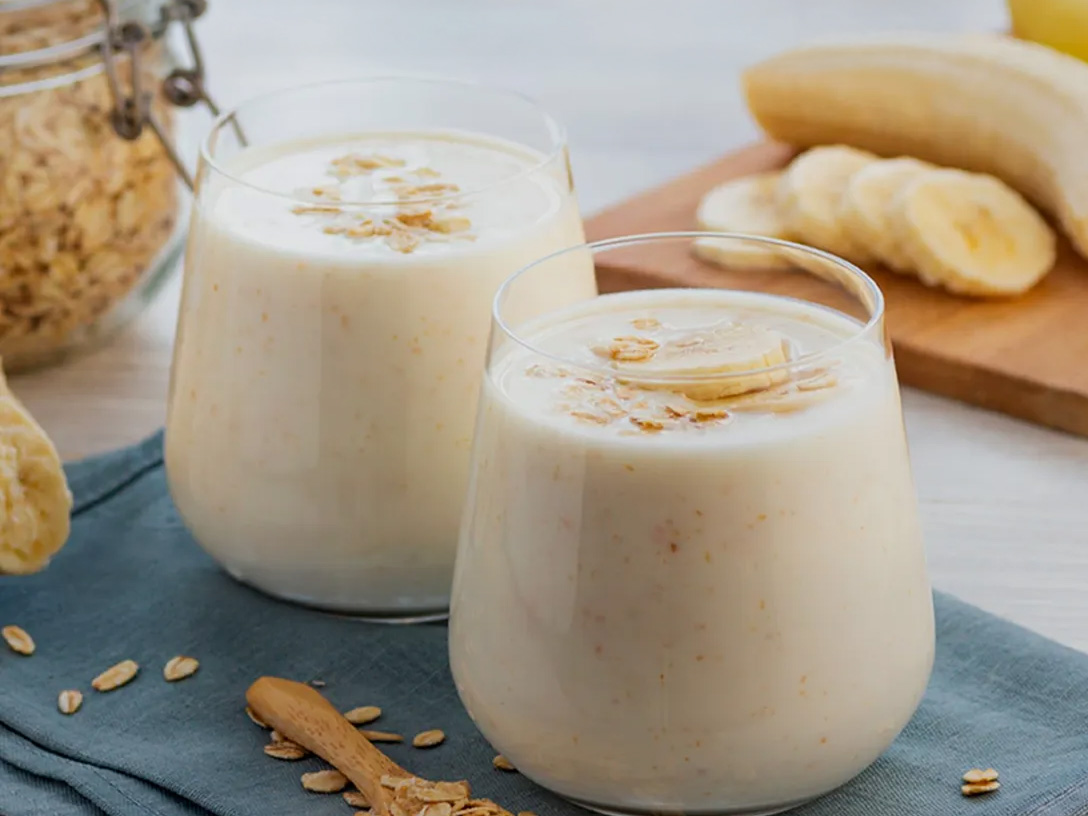 Dos vasos de licuado de plátano con avena, decorados con rodajas de plátano y avena espolvoreada, sobre una tela azul. Al fondo, se ve un frasco de avena, un plátano entero y rodajas de plátano en una tabla de madera. En la parte superior de la imagen, un texto dice "LICUADO DE PLÁTANO CON AVENA" y debajo "PREPÁRALO CON LECHE SANTA CLARA".
