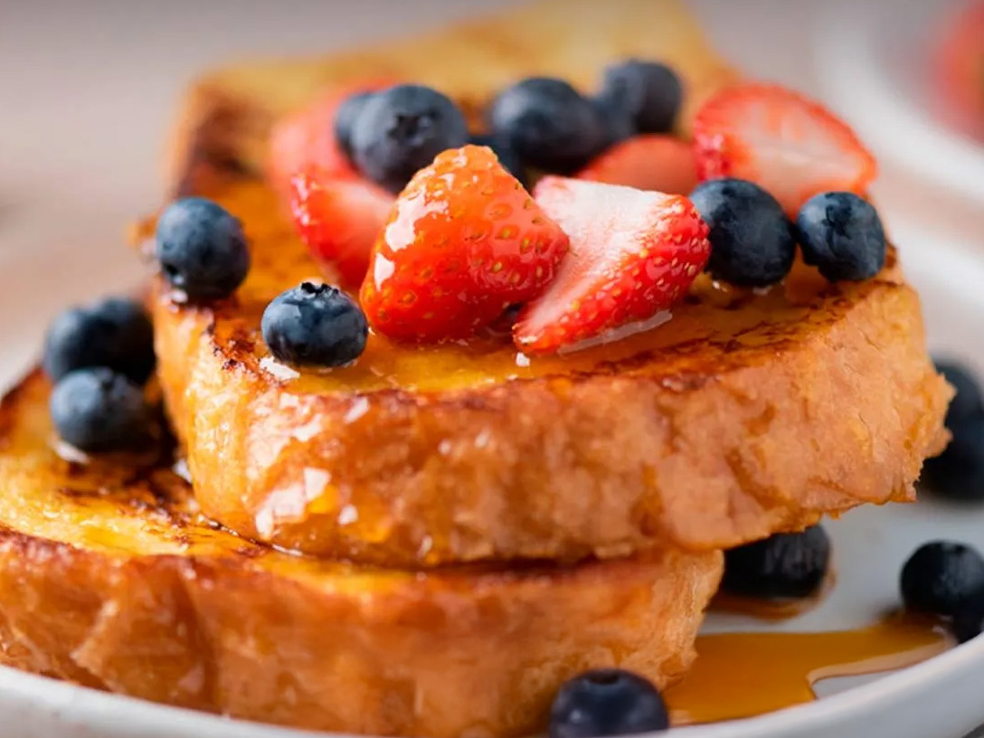 Dos rebanadas de tostadas francesas doradas apiladas en un plato blanco, cubiertas con arándanos frescos y fresas en rodajas, y bañadas en jarabe.