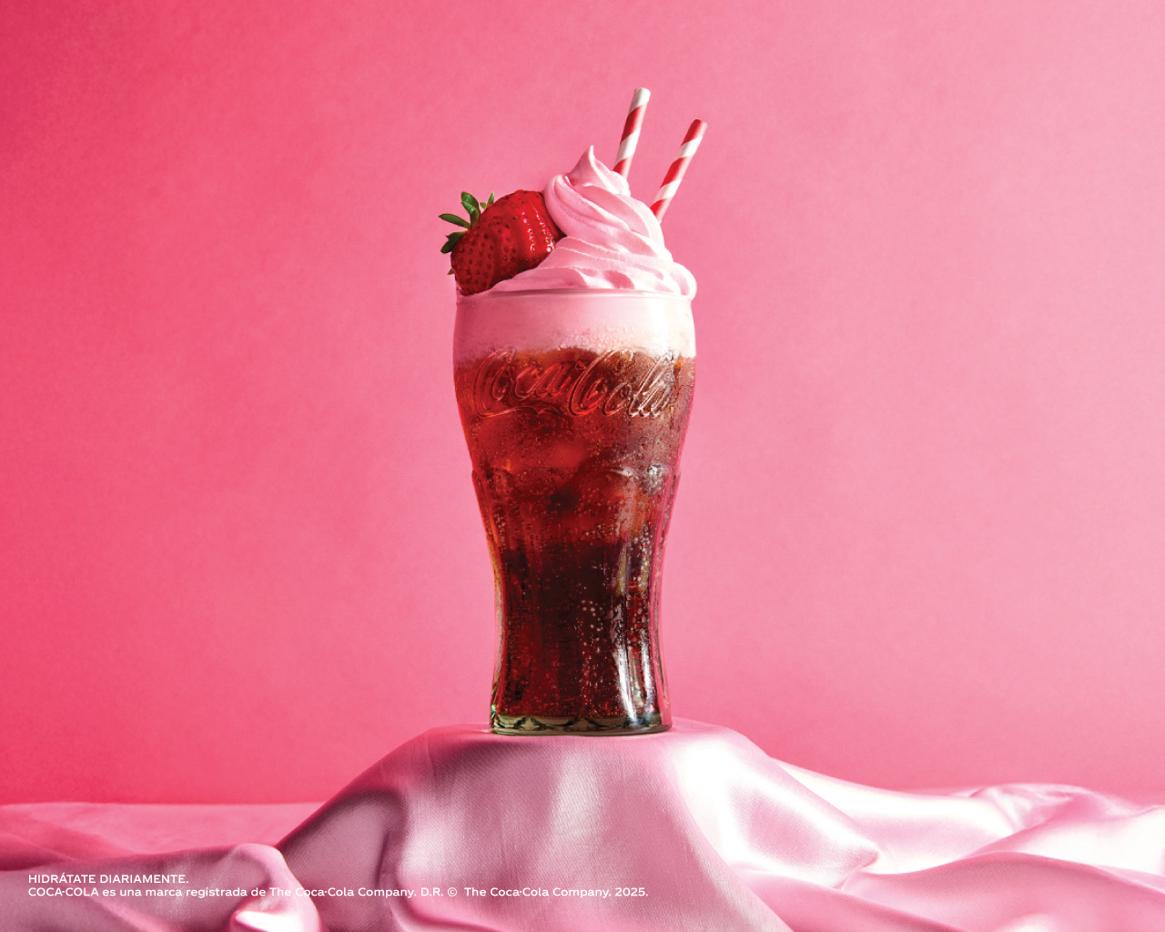 Bebida especial de Coca-Cola con crema batida y decoraciones de colores, presentada en vaso de vidrio sobre base decorativa.​