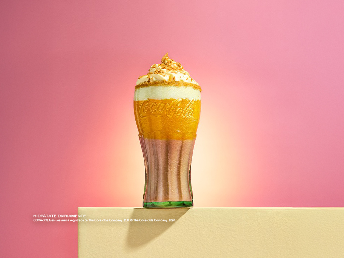 Bebida especial de Coca-Cola con crema batida y decoraciones de colores, presentada en vaso de vidrio sobre base decorativa