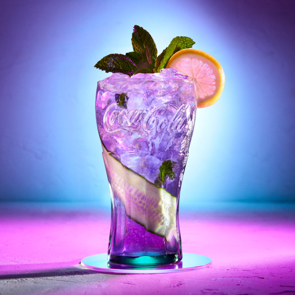 Bebida exclusiva servida en vaso de vidrio Coca-Cola con hielo, rodaja de limón y hojas de menta, sobre fondo iluminado en tonos morado y azul.​