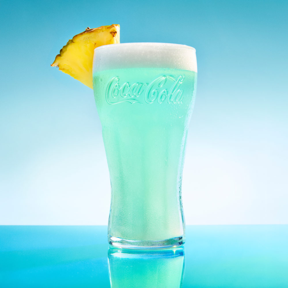Bebida servida en vaso de vidrio Coca-Cola con líquido color turquesa, espuma blanca en la parte superior y rodaja de piña como decoración, sobre fondo azul.​