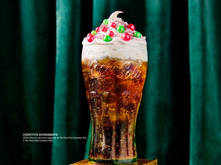 Bebida especial de Coca-Cola con crema batida y decoraciones de colores, presentada en vaso de vidrio sobre base decorativa
