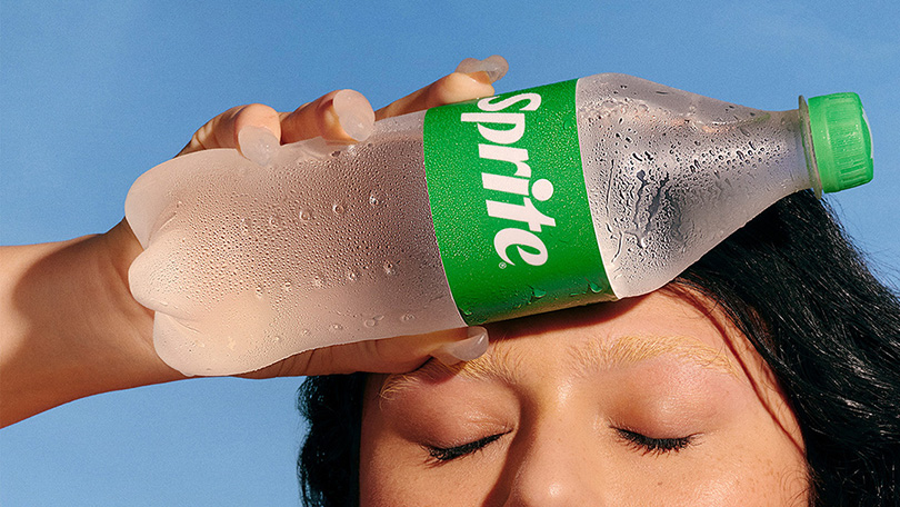 Mujer de cabello negro rizado bajo un cielo celeste con una botella de Sprite en su mano sosteniendola contra su frente