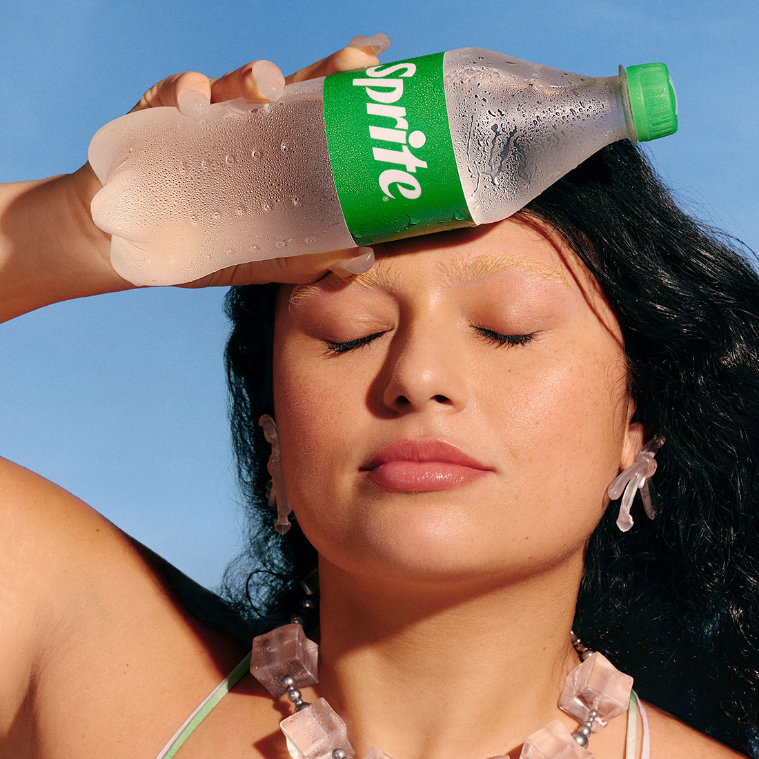 Mujer de cabello negro rizado bajo un cielo celeste con una botella de Sprite en su mano sosteniendola contra su frente