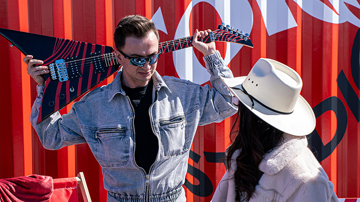 Un hombre sosteniendo una guitarra detrás de su cabeza, conversando con una mujer que lleva un sombrero de vaquero, con un vibrante fondo de Coke Studio.