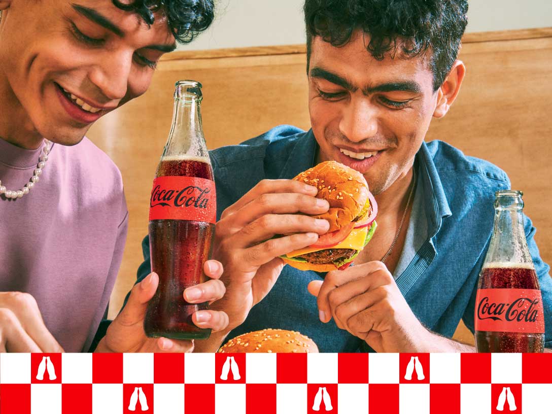 Dos jóvenes sonríen mientras disfrutan de hamburguesas y Coca-Cola en un ambiente relajado. En la parte inferior de la imagen, hay un patrón de cuadros rojos y blancos con siluetas de botellas.