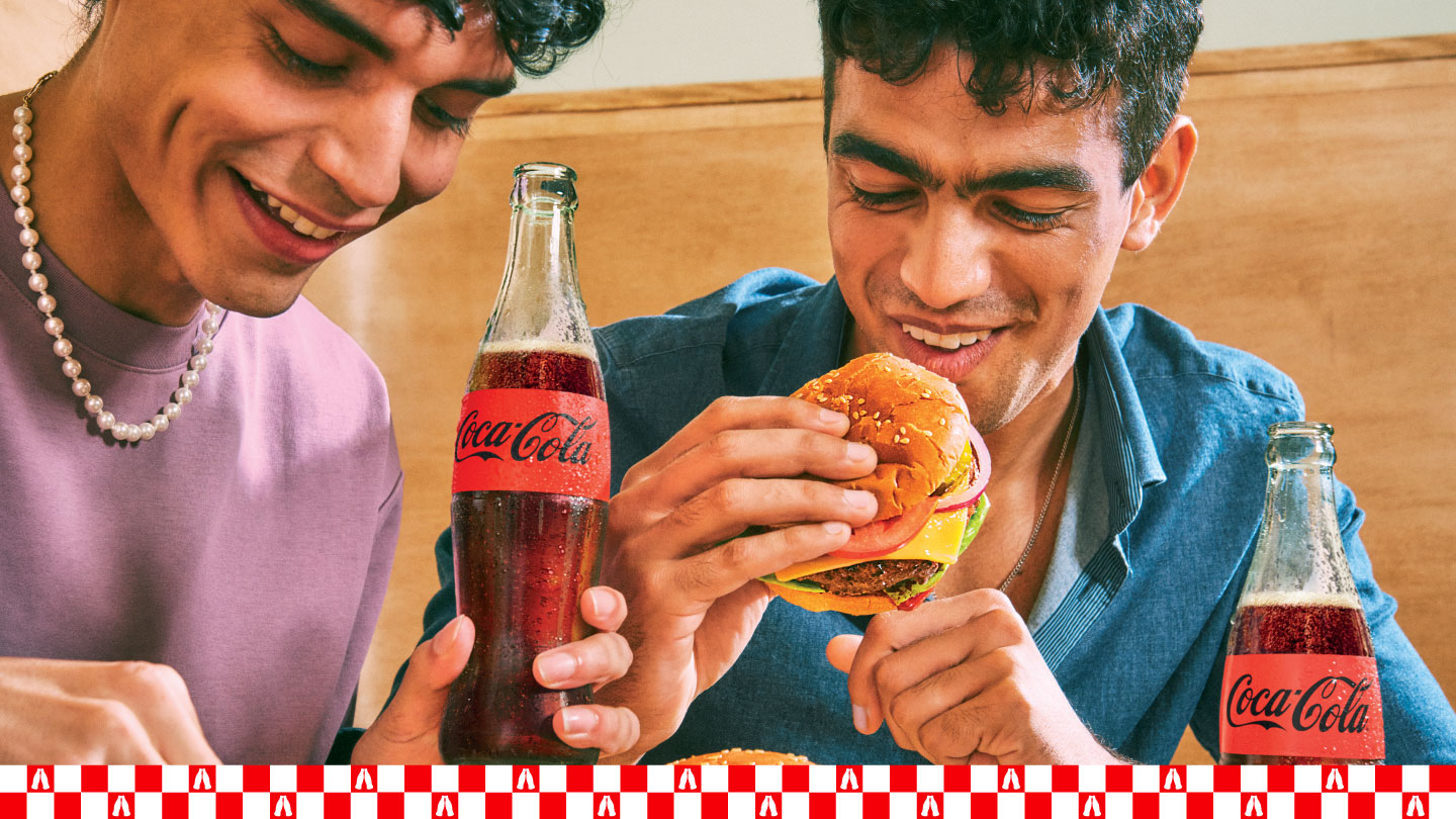 Dos jóvenes sonríen mientras disfrutan de hamburguesas y Coca-Cola en un ambiente relajado.