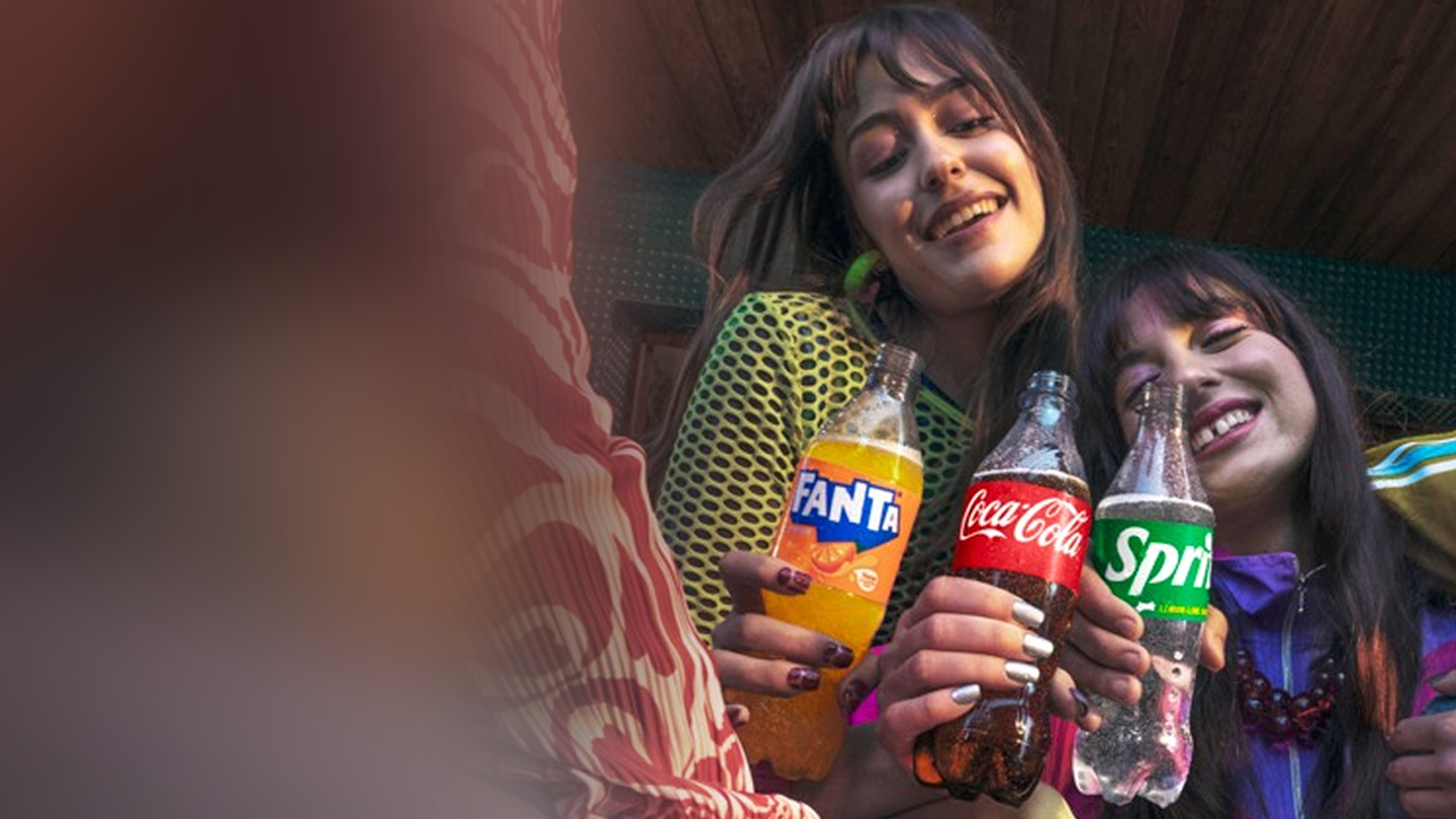 Nasmejana mlada ženska v turkiznem puloverju gleda v telefon in stoji ob kolesu – vabi k prijavi v digitalni svet Coca-Cola z nagradami in igrami.
