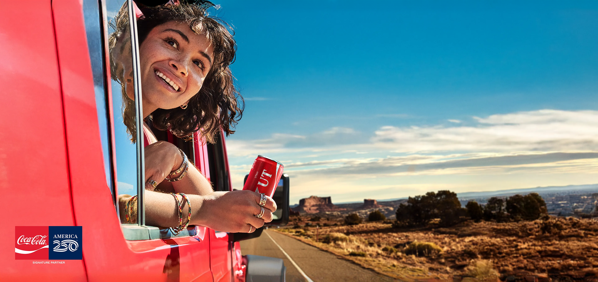 Smiling woman holding a Utah Coca-Cola can out a red car window on a desert road trip, with America 250 partner logo.