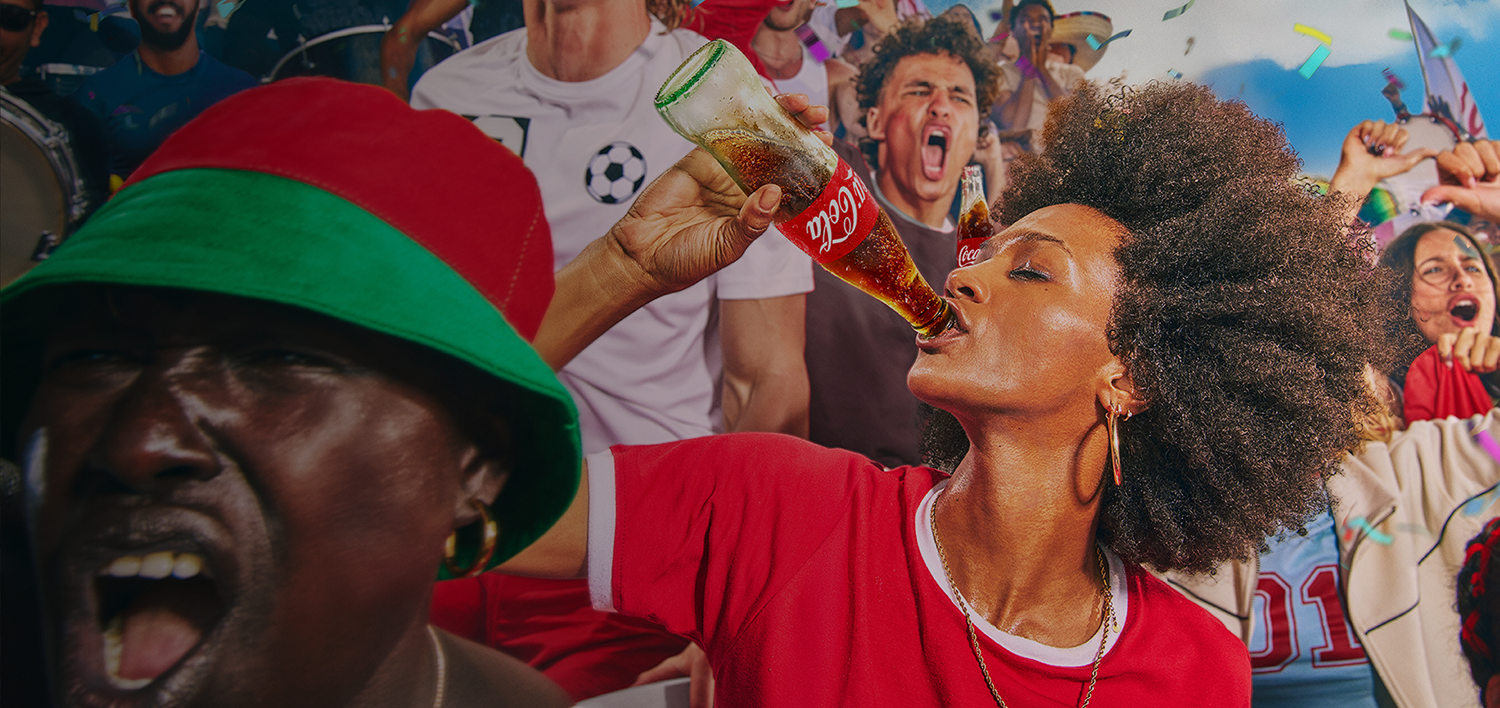 Soccer fan in red shirt drinking Coca‑Cola bottle in packed stadium as confetti falls, celebrating FIFA soccer.