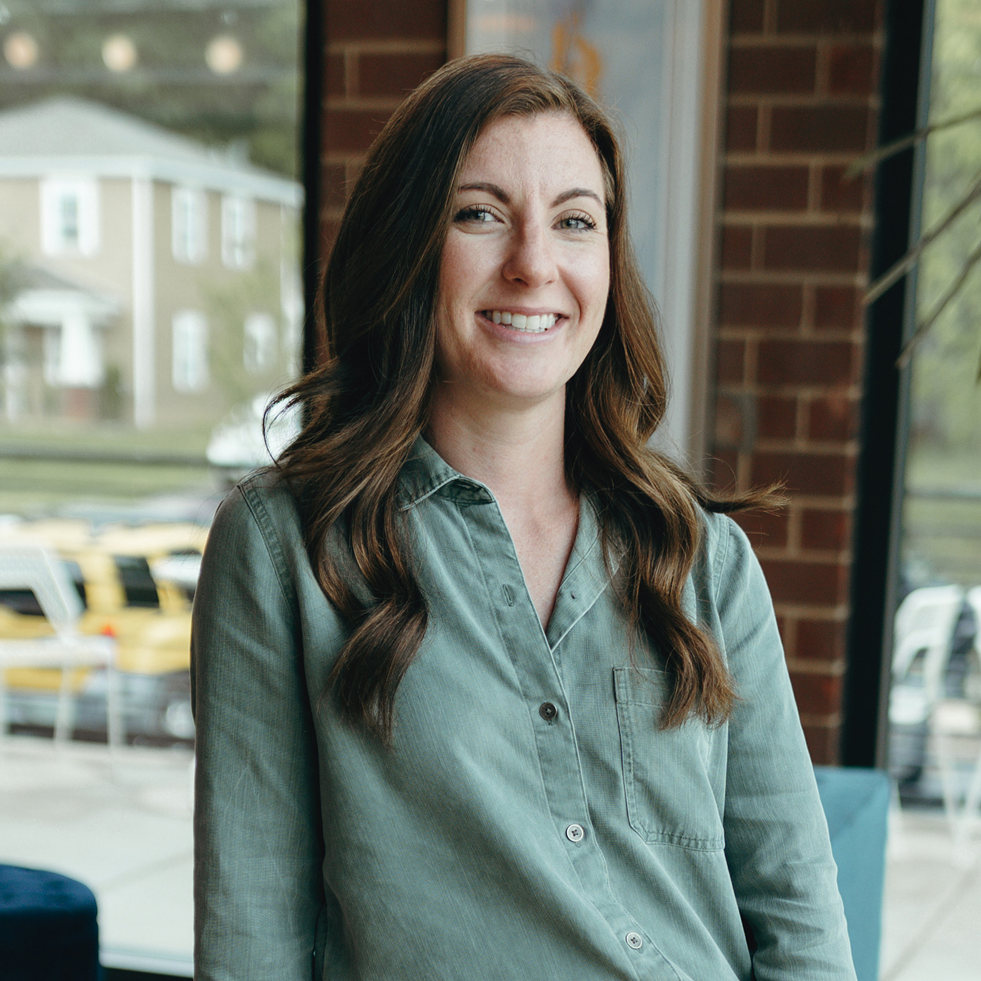 Mural artist with long wavy hair wearing green button‑up shirt standing indoors by large windows and brick wall.