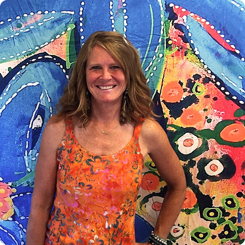 Artist wearing bright orange floral tank top standing in front of large abstract blue and coral painting in studio setting.