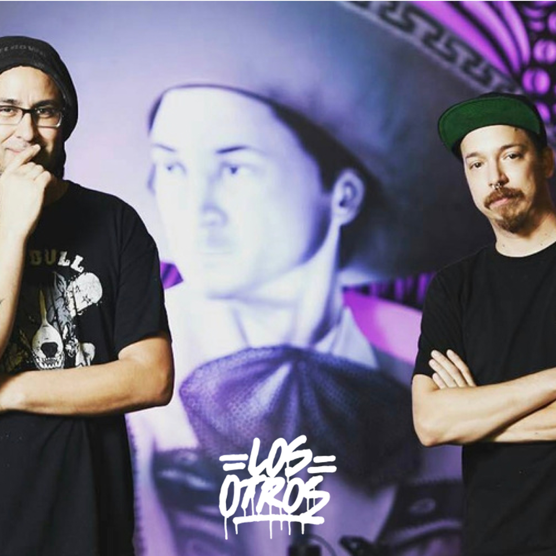 Profile shot of the mural artist duo, Los Otros, standing in front of one of their artworks with their logo in the foreground.