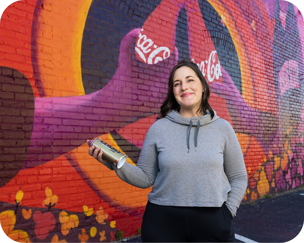 Artist Meg Wagler holding a spray paint can in front of her vibrant Coca‑Cola mural.