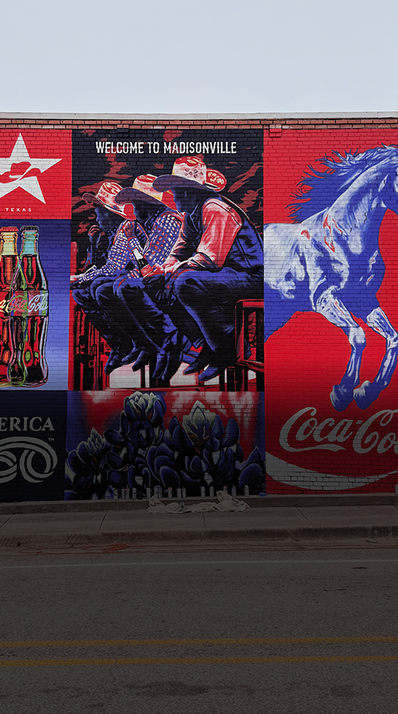 A vibrant red, white, and blue mural on a brick wall that reads "Welcome to Madisonville" with images of cowboys and Coca-Cola.