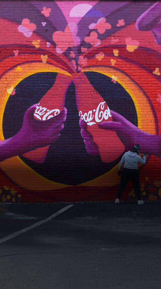 An artist paints a large, colorful mural of two hands toasting with Coca-Cola bottles as part of the Paint the Nation campaign.