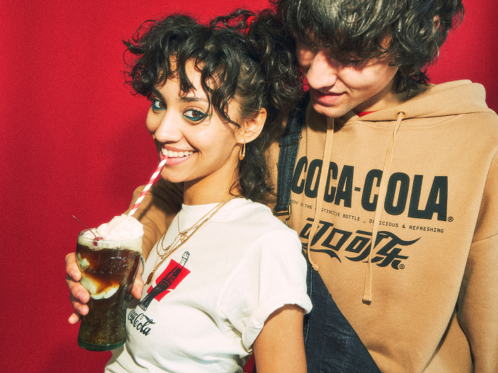 Two young people pose against a red background; one holds a glass of cola with a red-and-white striped straw, both wearing Coca-Cola logo clothing.