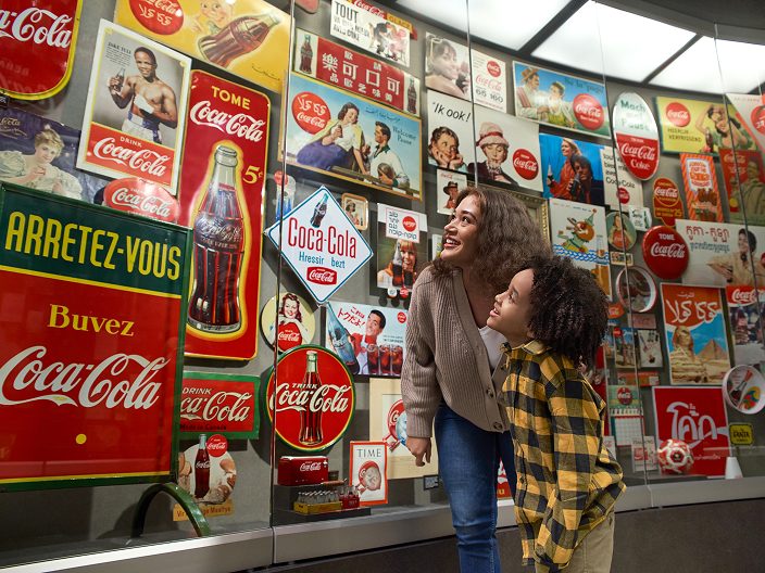 A woman and young child look up at a curved wall displaying vintage international Coca-Cola advertisements and memorabilia.